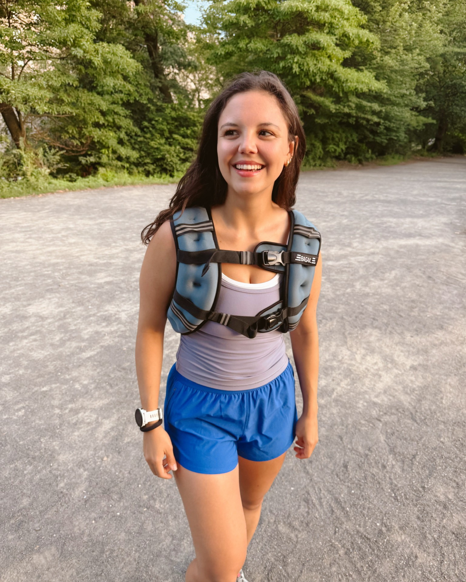 Woman walking outdoors on a gravel path, smiling confidently. She’s wearing a weighted vest over a fitted tank top and blue athletic shorts, with a smartwatch on her wrist. Trees and greenery line the background.